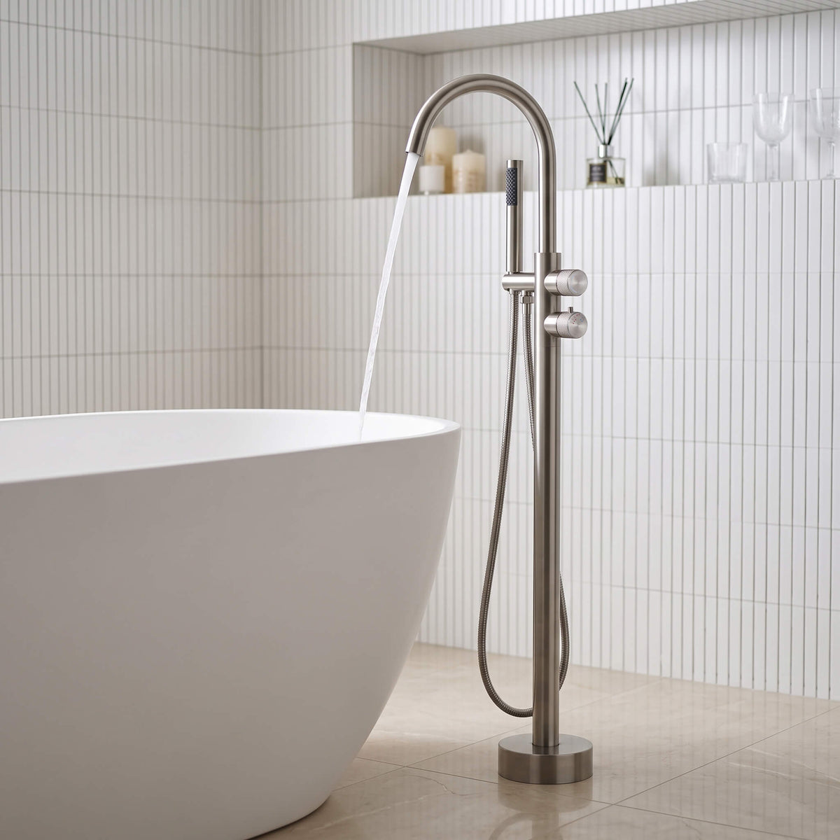 Modern freestanding bathtub faucet with hand shower in brushed nickel finish beside a white freestanding bathtub