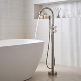 Modern freestanding bathtub faucet with hand shower in brushed nickel finish beside a white freestanding bathtub
